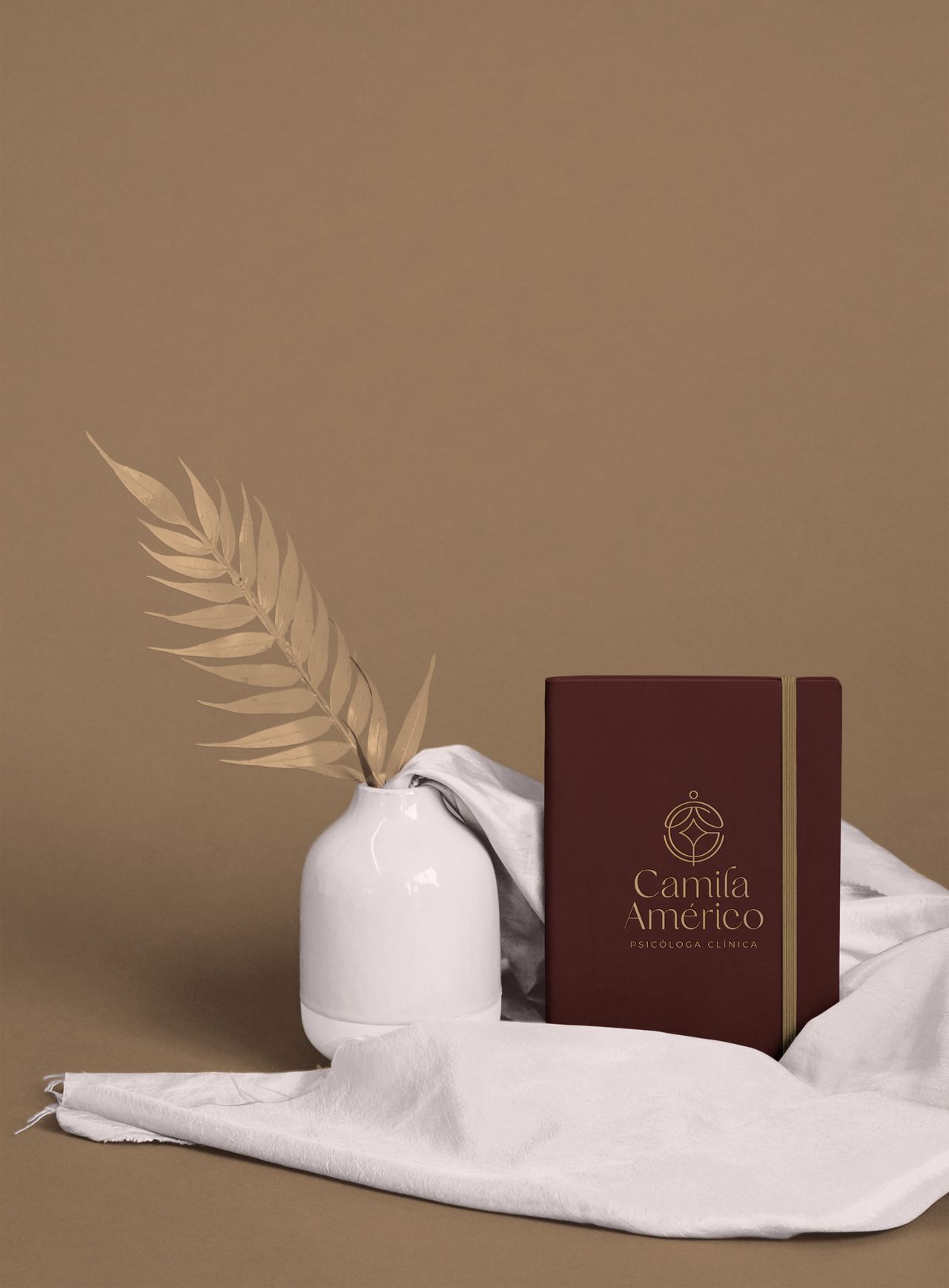 Caderno com a marca Camila Américo Psicóloga Clínica ao lado de um vaso branco com folha seca